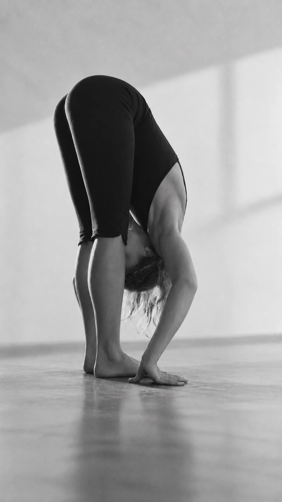 Black-and-white photograph: a standing forward fold, the hands flat on the concrete floor beside the feet, the hair falling towards the ground