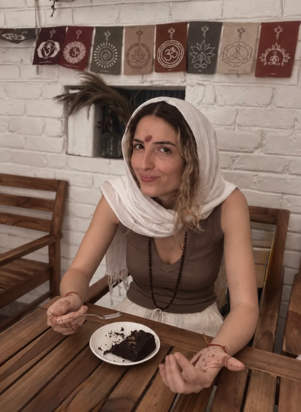 Irene at a wooden café table in Tapovan, Rishikesh, a white scarf over her hair, henna on her hands, smiling over a plate with a brownie